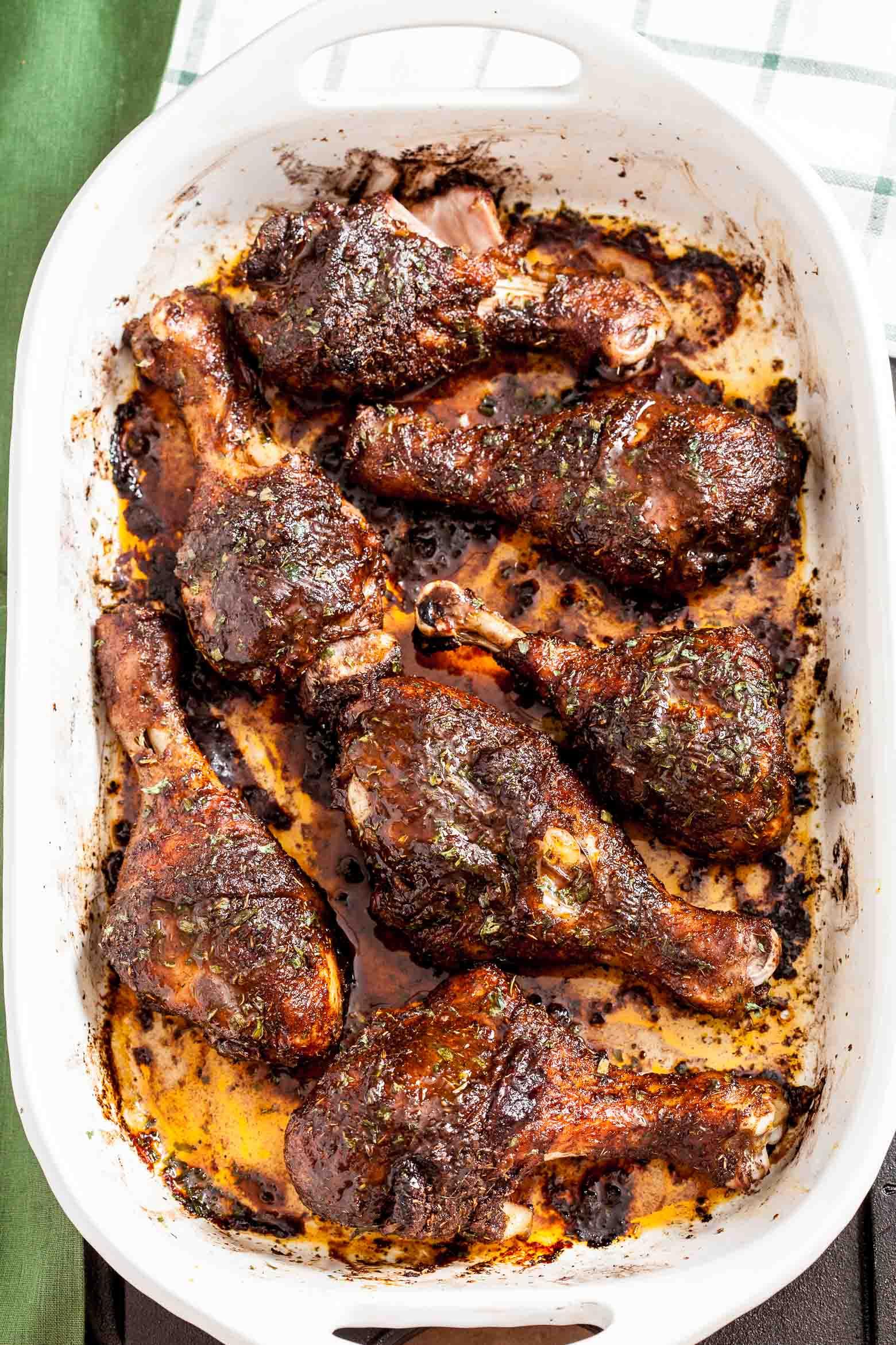Golden-brown Jamaican jerk chicken legs with caramelized spice crust on a baking sheet, garnished with fresh herbs and lime wedges