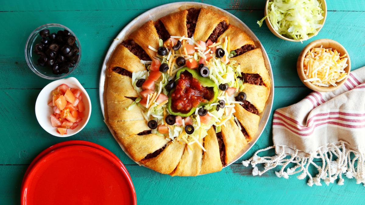 Golden-brown taco ring made with crescent rolls filled with seasoned ground beef, topped with fresh lettuce, tomatoes, and cheese, with salsa in the center