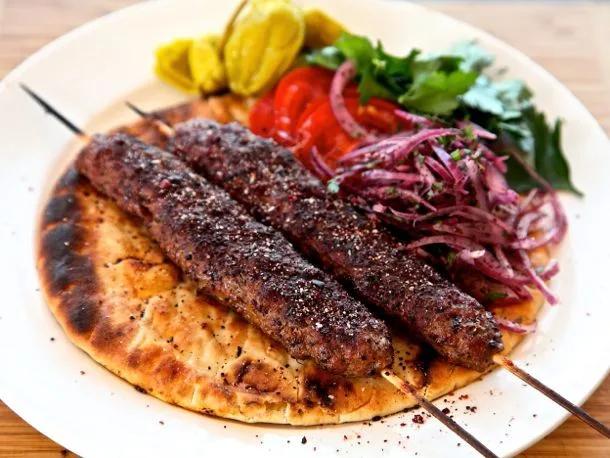 Golden charred Turkish Adana kebabs on skewers with fresh herbs, sumac onions, and warm pita bread on rustic wooden serving board