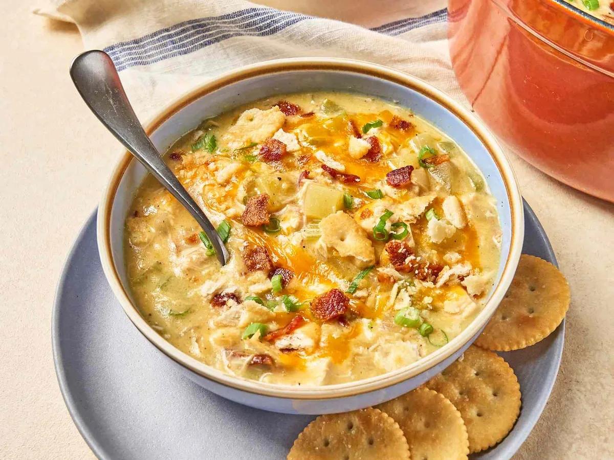 Creamy million dollar soup in a white bowl topped with bacon, cheddar cheese, green onions and crushed Ritz crackers