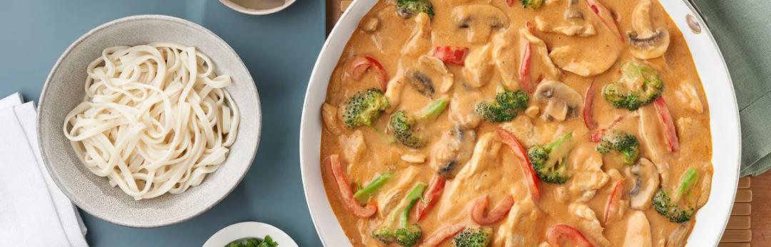 A bowl of curry chicken with thick udon noodles, red and yellow peppers, mushrooms and broccoli in a golden curry sauce